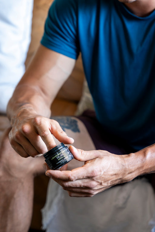A man in a blue shirt and black shorts with a tattoo on his leg using a CBD rub
